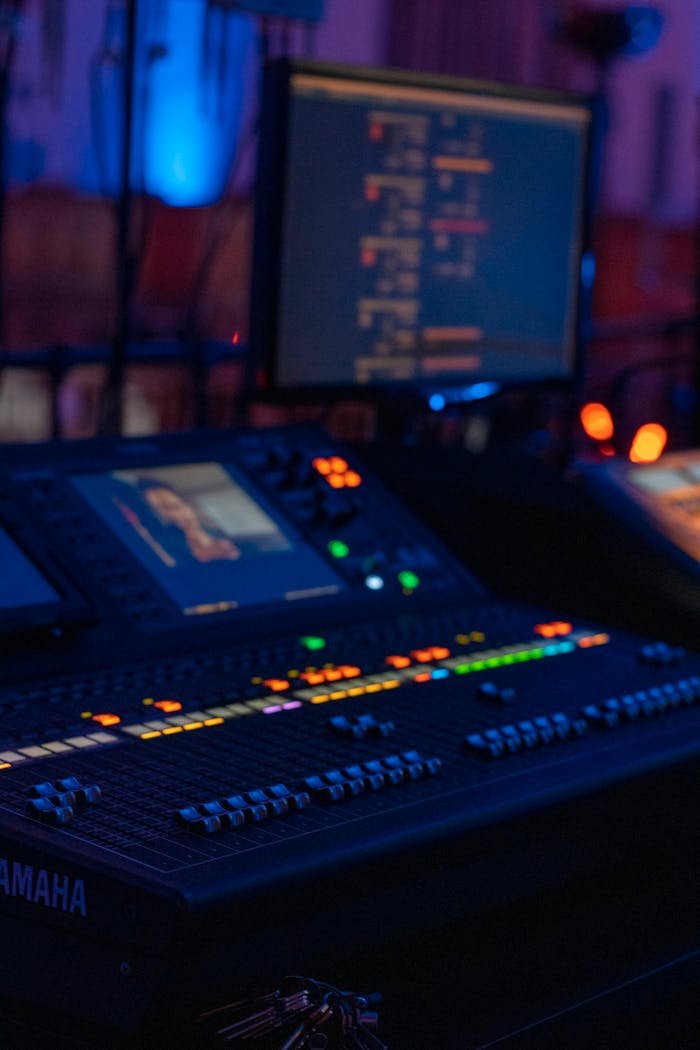 Close-up of a professional audio mixing console in a dimly lit music studio with monitors displaying audio software.